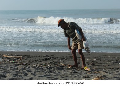 Yogyakarta, Indonesia, February 1, 2022 ; Selective Focus, A Traditional Fisherman Is Cleaning Up Trash On A Black Sandy Beach With Small Waves On A Sunny Day. Photo Is Noise