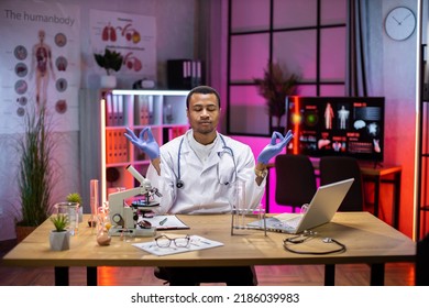 Yong African American Male Scientist Relaxing Practicing Yoga Wearing Lab Coat Working In Laboratory While Examining Biochemistry Sample In Test Tube And Scientific Instruments.