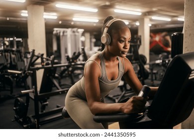 A yong african american sportswoman in shape with headphones doing workouts on a bicycle at gym. - Powered by Shutterstock