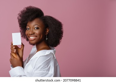 Yong beautiful happy smiling African American woman, model wearing elegant jewelry, classic shirt, holding small white box, posing in studio, on pink background. Copy, empty space for text