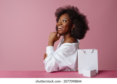 Yong Beautiful Happy Smiling African American Woman, Model Wearing Classic Shirt, Posing With  Small White Box, Shopping Paper Bag, In Studio, On Pink Background. Copy, Empty Space For Text