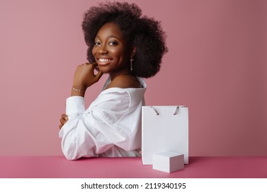 Yong Beautiful Happy Smiling Black  Woman, Model Wearing Classic Shirt, Jewelry, Posing With Small White Box, Shopping Paper Bag, In Studio, On Pink Background. Copy, Empty Space For Text
