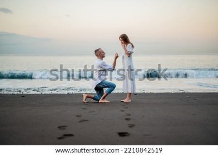 Yong man making a marriage proposal on a beach at sunset to a young beautiful woman. Standing on on knee.