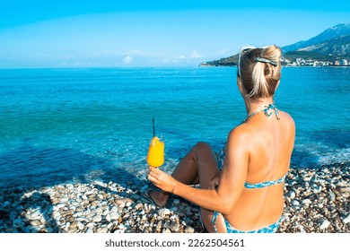 Yong woman sits on a clean pebble beach, holding a glass of orange fresh juice in her hand. Blue calm sea in morning. Summer holiday vacation concept. - Powered by Shutterstock