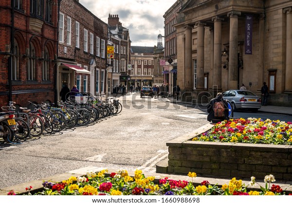 York,\
UK.  February 26, 2020.    Blake Street in York.  There is a flower\
bed in the foreground and a row of bicycles.  Historic buildings\
line the street and there is a cloudy sky\
above.