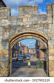 York, UK. February 26, 2021.  A View Down A Busy City Street Through An Ancient Archway Cut Into A Wall.  People Walk Down The Street And Cars Wait At A Traffic Light.