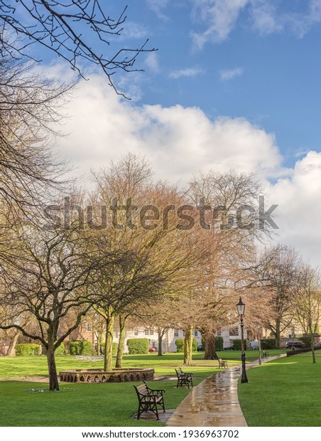 York, UK.  March 12, 2021.  York\
Minster gardens after a rain shower.  The path is wet and benches\
are one side with trees beyond. A cloudy sky is\
above.