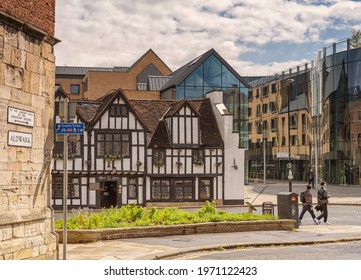 York, UK. May 7, 2021. The Ancient Black Swan, A Haunted Public House Standing Beside A Modern Glass Building. A Flower Bed In Is The Foreground And A Cloudy Sky Is Overhead.