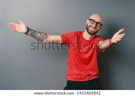 You are always welcome. Handsome young man with glasses, beard and tatooes, in red t-shirt gesturing and smiling while standing against grey background. Designer welcomes you.
