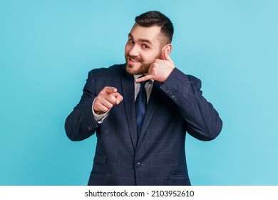 You contact us! Portrait of positive man with beard in dark suit making gesture with fingers dial my number or call me back and pointing at you. Indoor studio shot isolated on blue background.