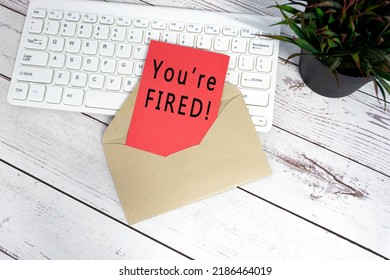 You Are Fired Text On Red Note Inside Brown Envelope With Keyboard And Potted Plant Background On Wooden Desk.