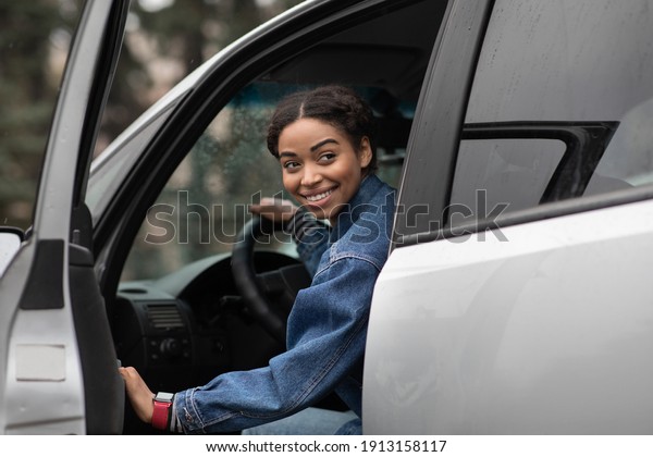 Are you going with me. Friendly smiling millennial\
african american female driver opens door and looks for fellow\
traveler, sitting in silver car or lady gets out of auto and looks\
back, copy space