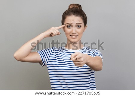 You are idiot. Woman wearing striped T-shirt showing stupid gesture and pointing to camera, blaming for insane plan, crazy idea, dumb suggestion. Indoor studio shot isolated on gray background.