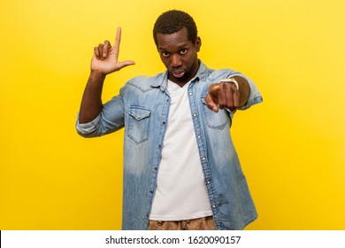 You are loser! Portrait of serious bossy man in denim casual shirt pointing at camera and showing lame or loser gesture, mocking your failures. indoor studio shot isolated on yellow background