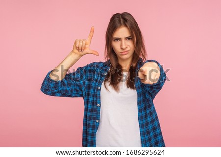 You lost! Portrait of girl in checkered shirt showing L sign with fingers, teasing you are loser and pointing to camera, accusing blaming for failure. indoor studio shot isolated on pink background