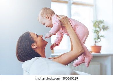 You Are My Treasure! Side View Of Cheerful Beautiful Young Woman Holding Baby Girl In Her Hands And Looking At Her With Love While Sitting On The Couch At Home