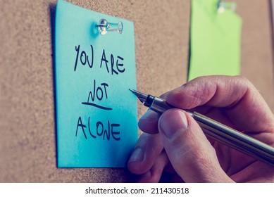 You Are Not Alone - man writing an inspirational message on a blue sticky note pinned to a cork board with a fountain pen.