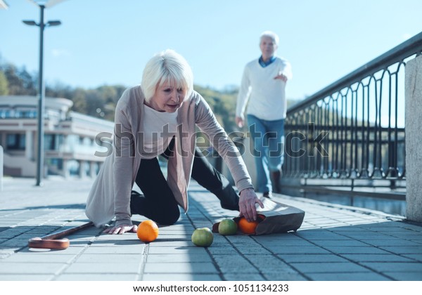 Are\
you ok. Selective focus on a scared senior lady standing on her\
knee and trying to pick up her groceries after falling down while\
her worried husband running to her in the\
background.