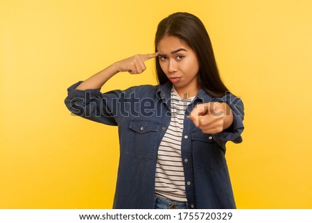 You are out of mind! Portrait of girl in denim shirt showing stupid gesture with finger near head and blaming fool for dumb insane plan, crazy idea. indoor studio shot isolated on yellow background