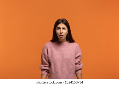Are you seriously. Stupefied attractive woman in pink sweater looks with astonishment into camera, being amazed with news. Expresses surprisement, doesn't believe his eyes. Isolated on orange wall