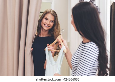 You should try on this dress! Beautiful young woman taking dress on hanger from seller while standing in fitting room at the store
