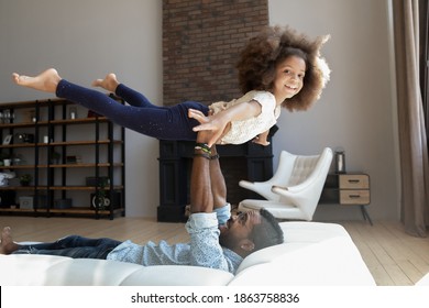 You Are So Strong, Daddy! Excited Active Little Black Preteen Girl Playing With Millennial Dad Flying In Air Stretching Arms And Legs While Father Lying On Sofa Holding Daughter In Arms Lifting Her Up