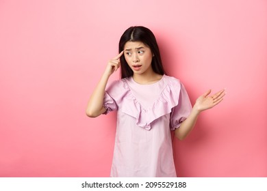 Are You Stupid. Shocked Asian Woman Pointing At Head And Complaining, Scolding Someone Crazy Or Strange, Look Aside, Standing Against Pink Background