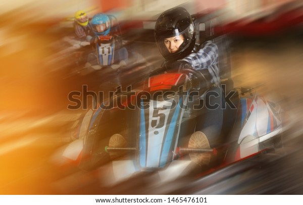 Youn woman in helmet driving car for karting\
with other people in sport club\
indoor