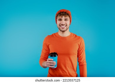 Yound attractive happy man in orange hat and t-shirt holds paper cup with coffee tea on blue background. High quality photo