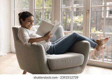 Young 25s smiling Indian woman reading novel book relaxing alone on comfort armchair in cozy room, spend leisure enjoy favourite literature bought in bookshop. Booklover pastime at home, hobby concept