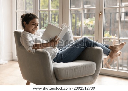 Young 25s smiling Indian woman reading novel book relaxing alone on comfort armchair in cozy room, spend leisure enjoy favourite literature bought in bookshop. Booklover pastime at home, hobby concept