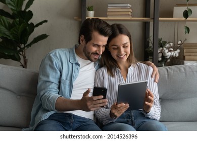 Young 30s couple sit on sofa use diverse gadgets holding smart phone and tablet device. Husband show explains to wife new mobile apps, comparing applications enjoy modern tech usage on weekend at home - Powered by Shutterstock