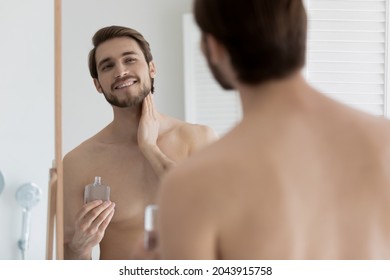 Young 30s handsome shirtless man looking in mirror applying to skin aftershave lotion, standing in bathroom do morning skincare routine. After shaving beauty skin care moisturizers products concept