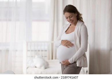 Young 30s happy pregnant woman in casual clothes embracing caressing her big belly, standing pose in light nursery at modern cozy home in last stages of pregnancy. Expectant mother, motherhood - Powered by Shutterstock