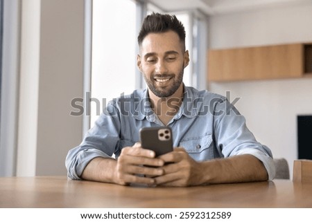 Young 30s Hispanic man using smartphone seated at table at home, smile while lead chat with girlfriend, using e-dating services mobile application, making call or buying goods though marketplace app