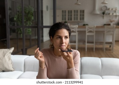 Young 30s Hispanic woman sit on sofa at home holding smart phone recording leaving audio message to friend, talking on speaker phone. Modern ai tech, advanced user, share voicemail activity concept