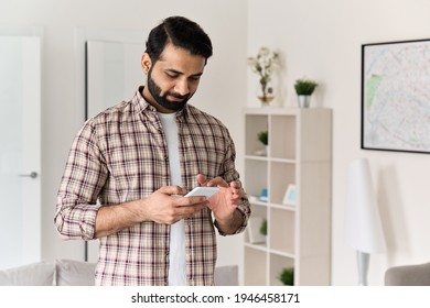 Young 30s Indian Man Holding Smartphone Tech Device Using Cell Phone Apps At Home. Bearded Ethnic Guy Texting Messages Looking At Smart Phone Checking Social Media, Ordering Online Or Browsing.