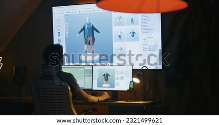 Young 3D designer creates video game character or clothes, works remotely from home in the evening on computer and big digital screen with professional software interface for 3D modeling and design.