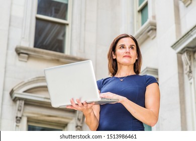 Young 40 Years Old Native American Businesswoman Working In New York City, Wearing Short Sleeve Dress, Holding Laptop Computer, Standing Outside Office Building With Windows, Looking Up, Thinking.
