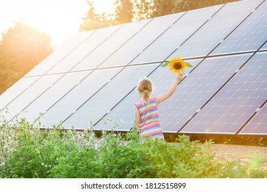 Young 6 Year Old Blonde Girl Child Standing In Front Of Small Solar Panel Farm In Countryside. Renewable Energy Concept. Sun Lens Flare.