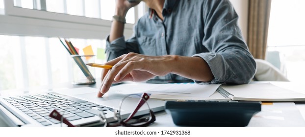 Young Accountant Hand Holding Smartphone To Call Marketing Consultants And Using Laptop Computer To Analyze Sales Growth In The Global Workplace Market. Accounting Concept