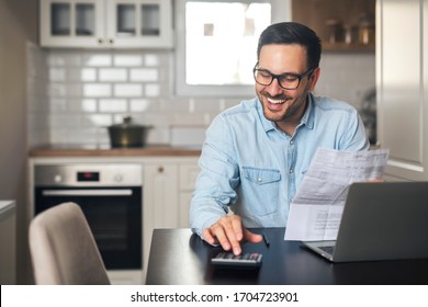 A young accountant sits in front of a laptop at home and holds a piece of paper while counting on a calculator. - Shutterstock ID 1704723901