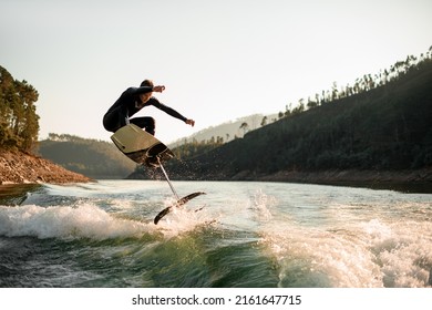 young active man in black wetsuit jump with foil wakeboard over splashing wave. Water sports activity.