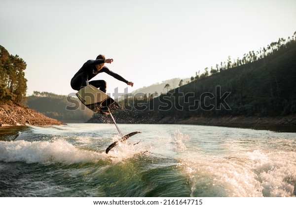 young active man in black\
wetsuit jump with foil wakeboard over splashing wave. Water sports\
activity.