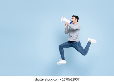 Young active man jumping and shouting on megaphone isolated on light blue background with copy space
