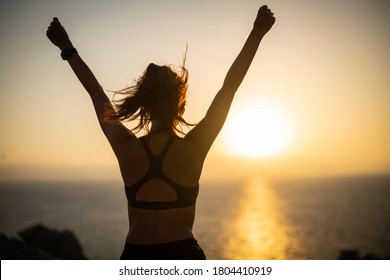 An young active sporty athlete woman with firm fit body is exulting of happiness satisfied with her achievement after running and jogging workout on a top of rock with seascape at sunset.