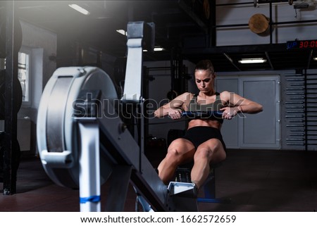 Young active strong fit sweaty powerful attractive muscular woman with big muscles doing hard core row heavy crossfit training workout on indoor rower at the gym real people exercise