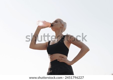 A young active woman with tattoos takes a break to drink water from a pink bottle during her outdoor fitness routine.