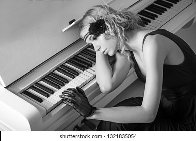 Young actress lying on piano overwhelmed with memories. Sad blond girl leaning on keyboard. Roaring twenties fashion concept.