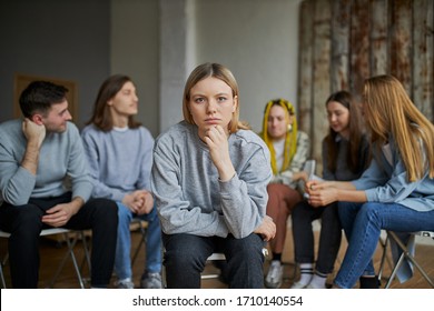 Young Addict Seriously Look At Camera, Sit Isolated In Room With Group Of People Having Conversation In The Background. Sick Female Need Help